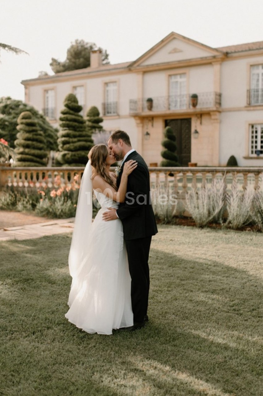 Un couple en tenue de mariage s'embrasse dans un jardin devant une grande maison élégante.