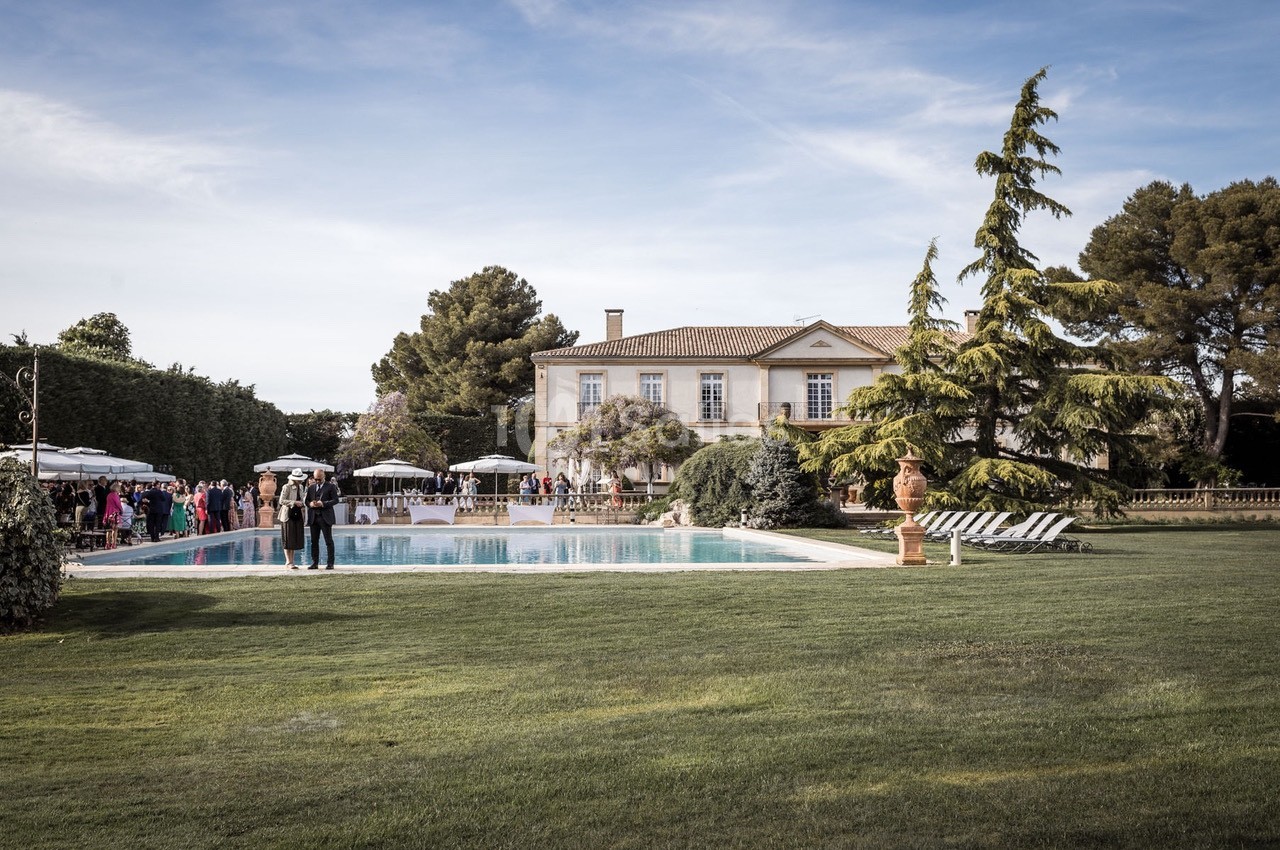 Grande maison avec piscine entourée de chaises longues, pelouse verdoyante et invités rassemblés sous des parasols.