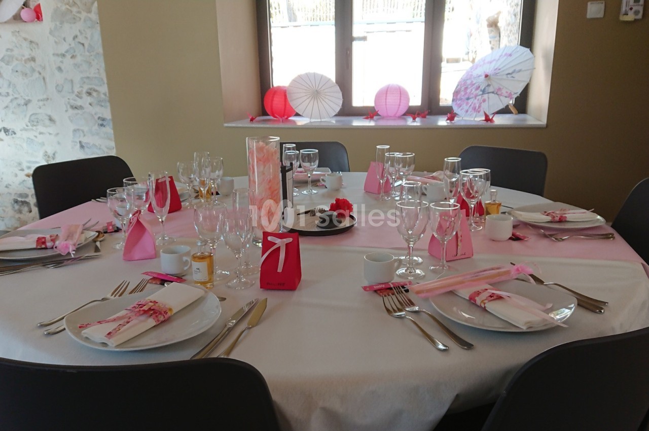 Table ronde décorée pour un événement, avec nappes blanches, serviettes roses, vaisselle et décorations élégantes.