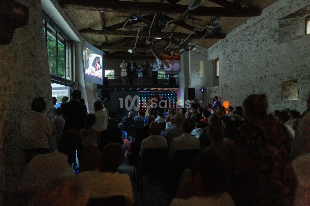Salle en pierre avec des invités assis, regardant une projection et une scène éclairée au fond.