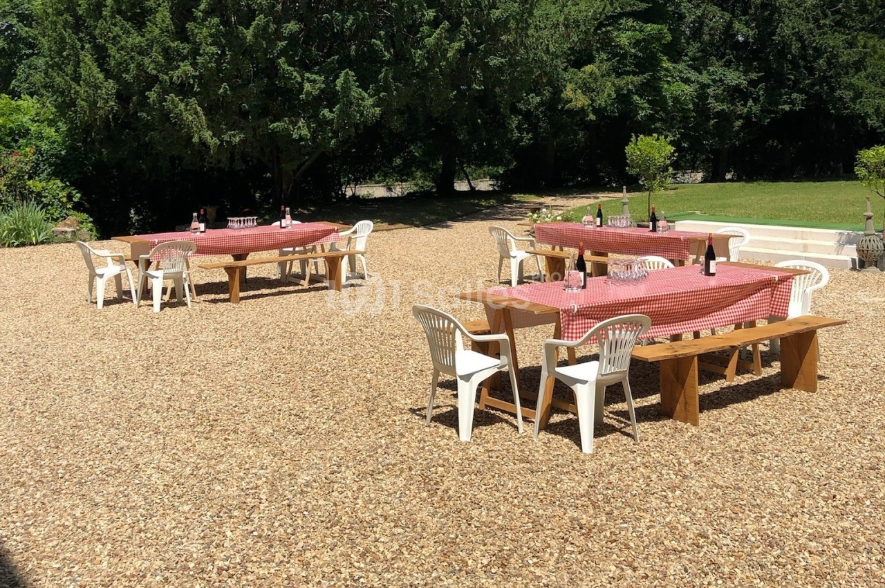 Tables en bois avec nappes rouges et chaises blanches disposées sur un sol gravillonné, entourées de verdure.