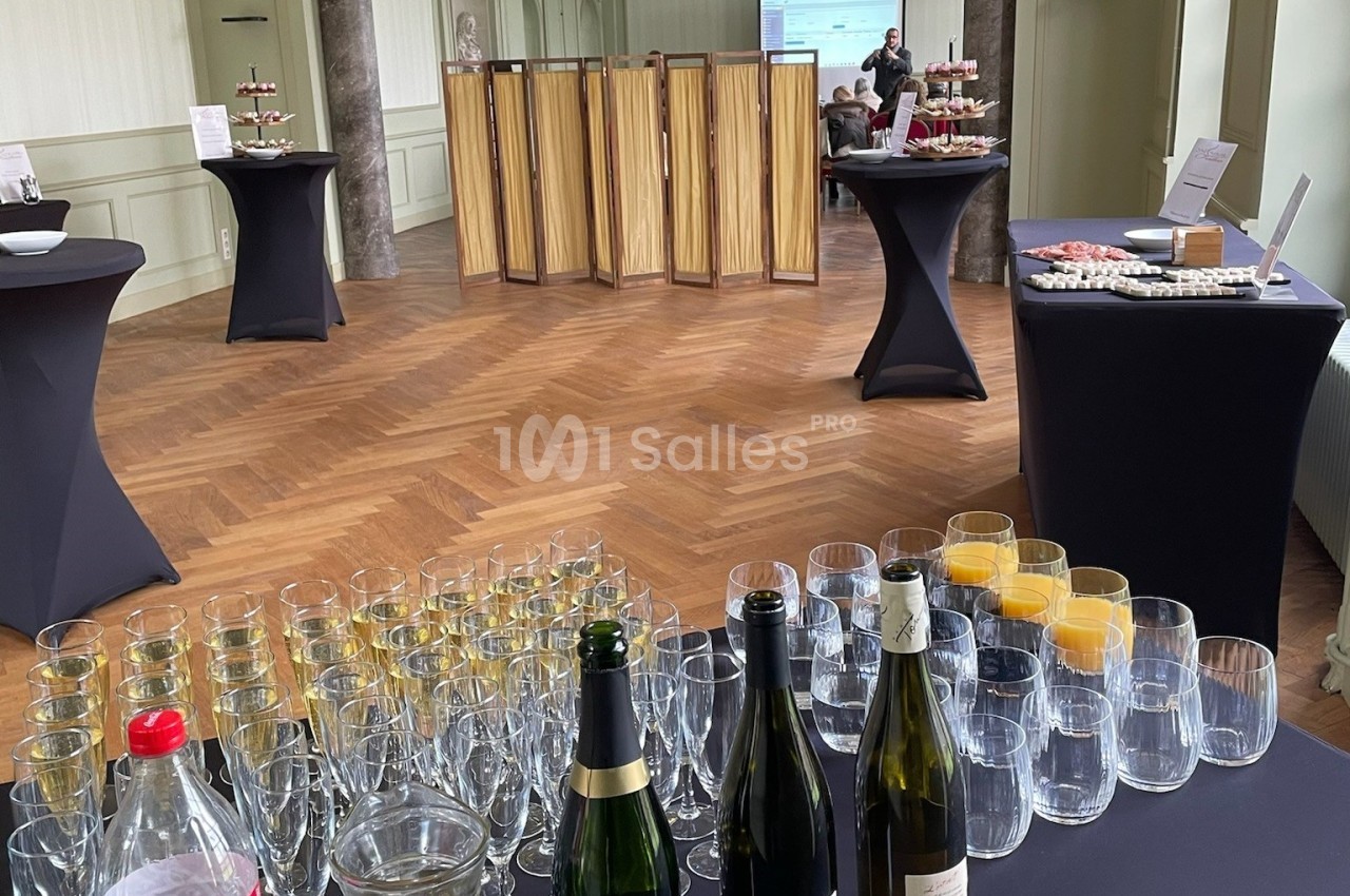 Salle avec parquet, tables hautes noires, boissons, verres et buffet disposés pour un événement ou une réception.