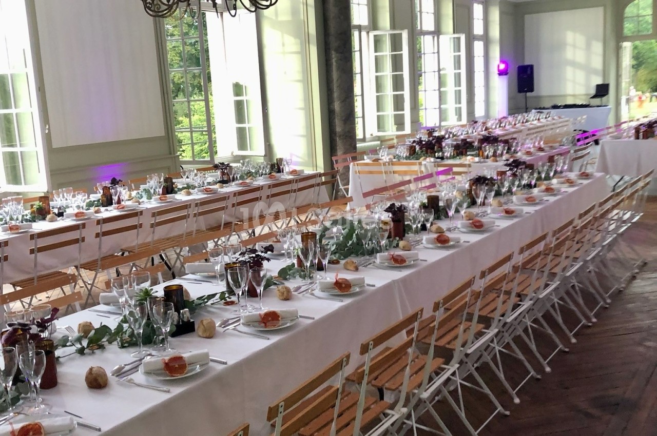 Salle de réception lumineuse avec de longues tables décorées de nappes blanches, vaisselle et éléments végétaux.
