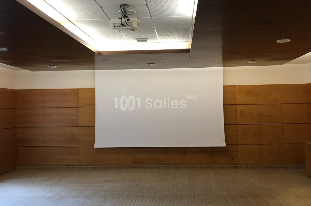 Écran de projection blanc déroulé dans une salle de conférence avec murs en bois et plafond équipé d'un projecteur.