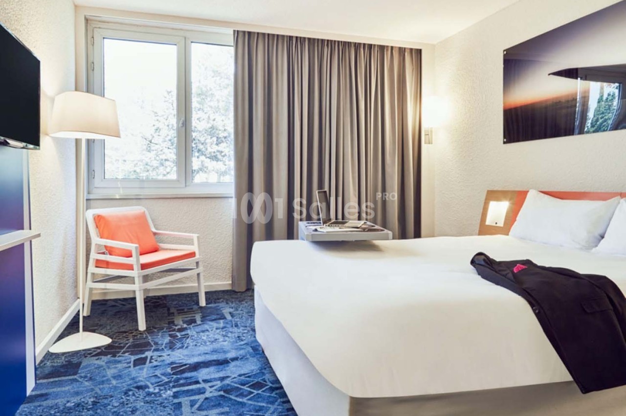 Chambre d'hôtel lumineuse avec un lit double, un fauteuil, une télévision, une fenêtre et des rideaux gris.