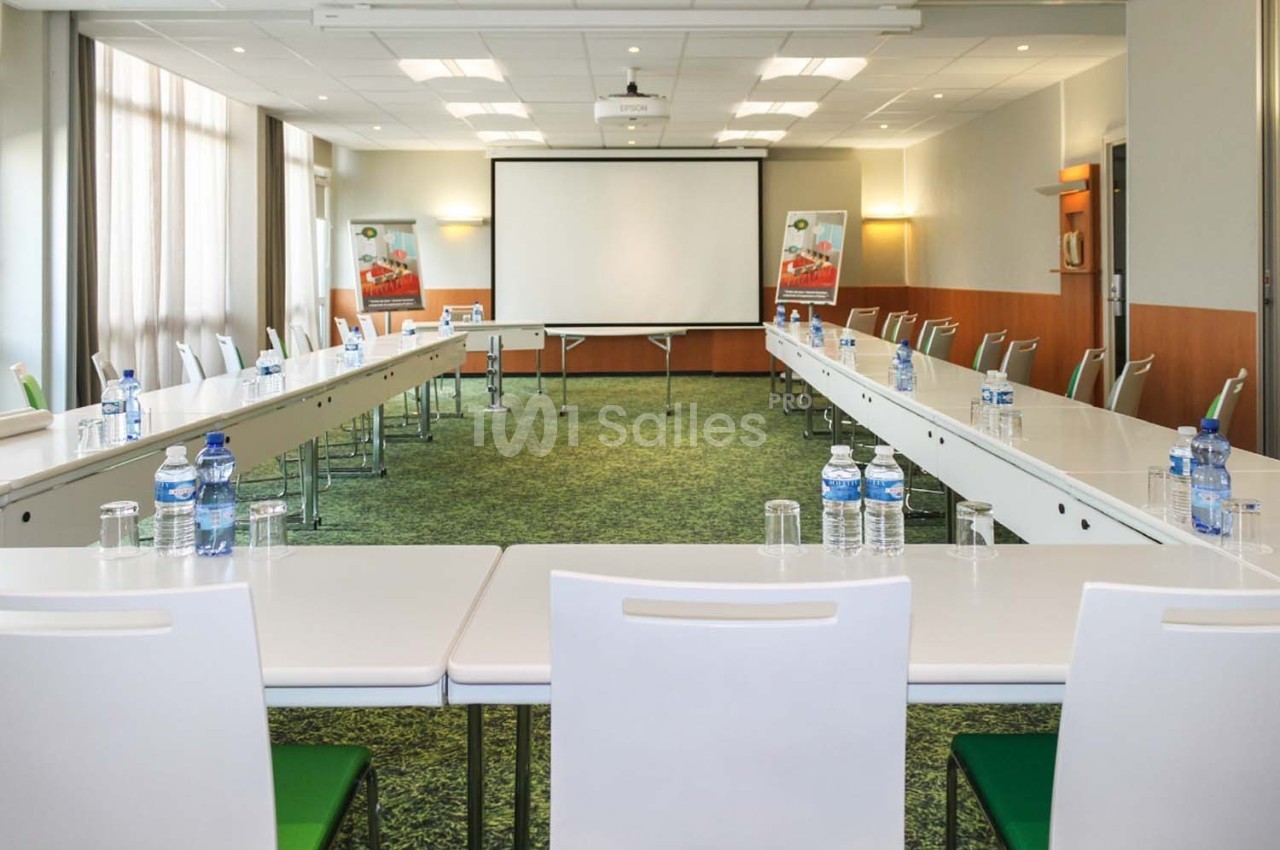 Salle de réunion lumineuse avec tables disposées en U, chaises blanches, écran de projection et bouteilles d'eau.