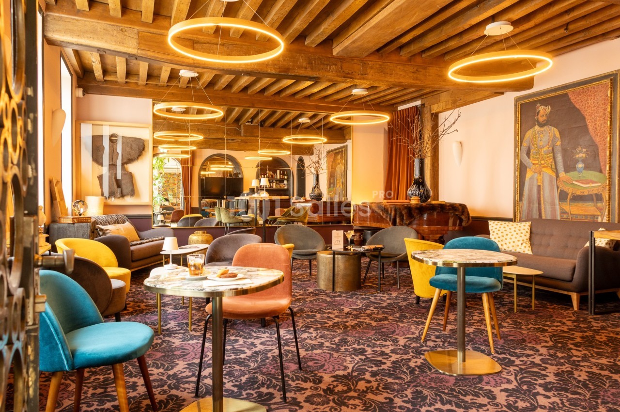 Salon chaleureux avec mobilier coloré, tables basses, éclairage suspendu circulaire et poutres apparentes au plafond.