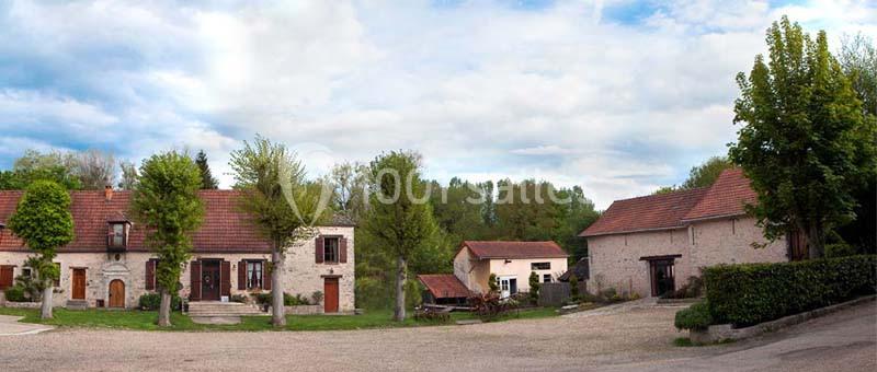 Bâtiments en pierre avec toits en tuiles rouges entourés d'arbres et d'une cour dégagée sous un ciel partiellement nuageux.