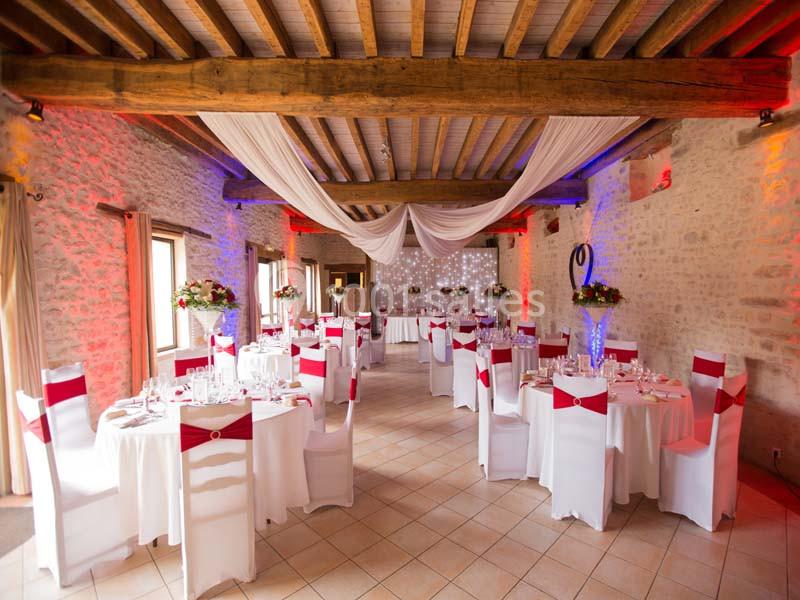 Salle de réception décorée avec des tables rondes, nappes blanches, chaises ornées de rubans rouges et éclairage chaleureux.
