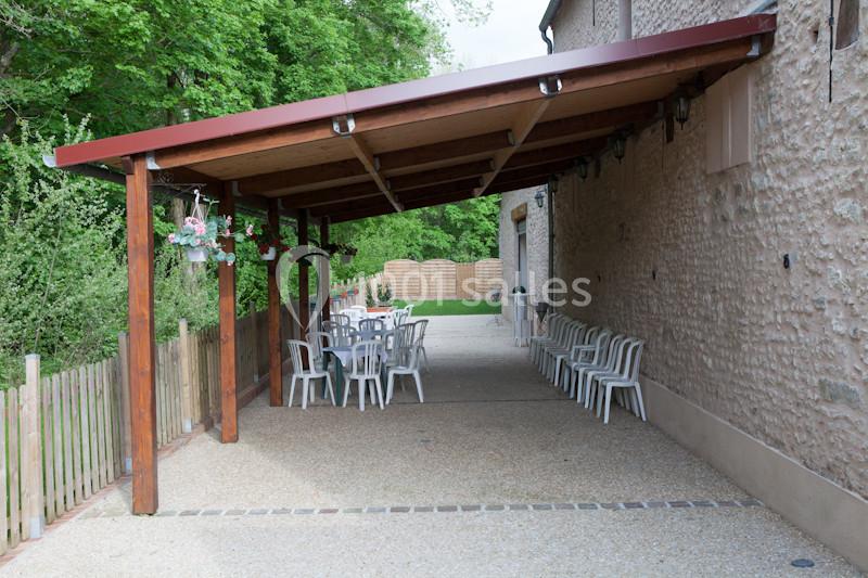 Abri en bois avec tables et chaises blanches, situé à côté d'un bâtiment en pierre, entouré de verdure.