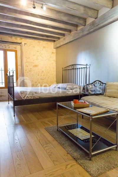 Chambre avec lit double, lit simple, murs en pierre, poutres apparentes et table basse en métal sur parquet.