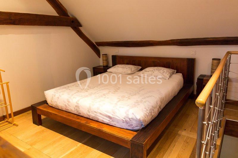 Lit double avec draps blancs dans une chambre mansardée, parquet en bois et poutres apparentes.