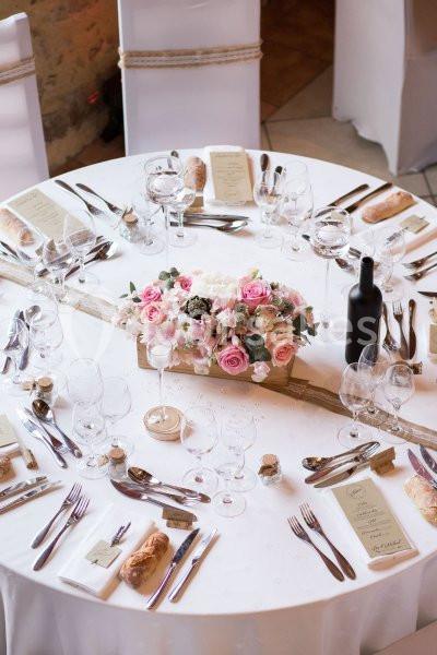 Table de réception élégamment dressée avec nappes blanches, couverts, menus, fleurs roses et bouteille de vin.