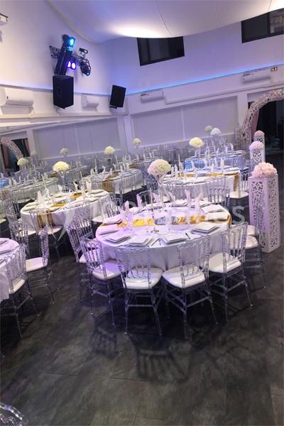 Salle de réception décorée avec des tables rondes, nappes blanches, chaises transparentes et centres de table fleuris.