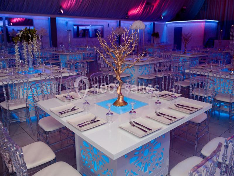 Salle de réception décorée avec des tables blanches ornées de centres de table dorés et éclairage coloré.