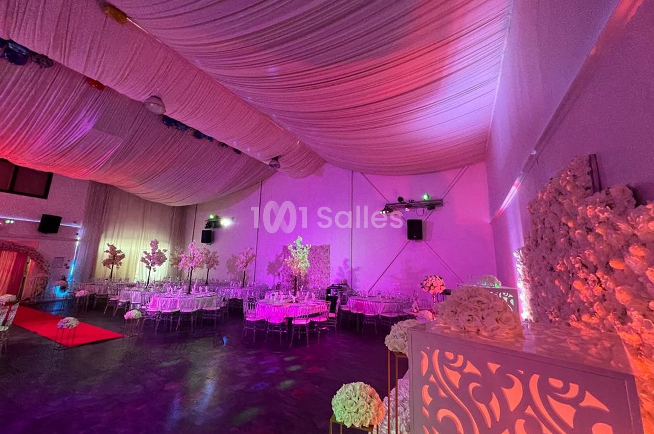 Salle de réception décorée avec des fleurs blanches, éclairages roses et violets, et tables dressées.
