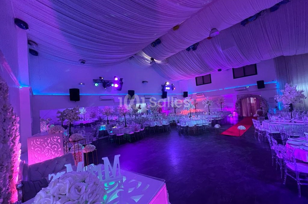 Salle de réception décorée avec des tables dressées, éclairages colorés et plafond drapé de tissu blanc.