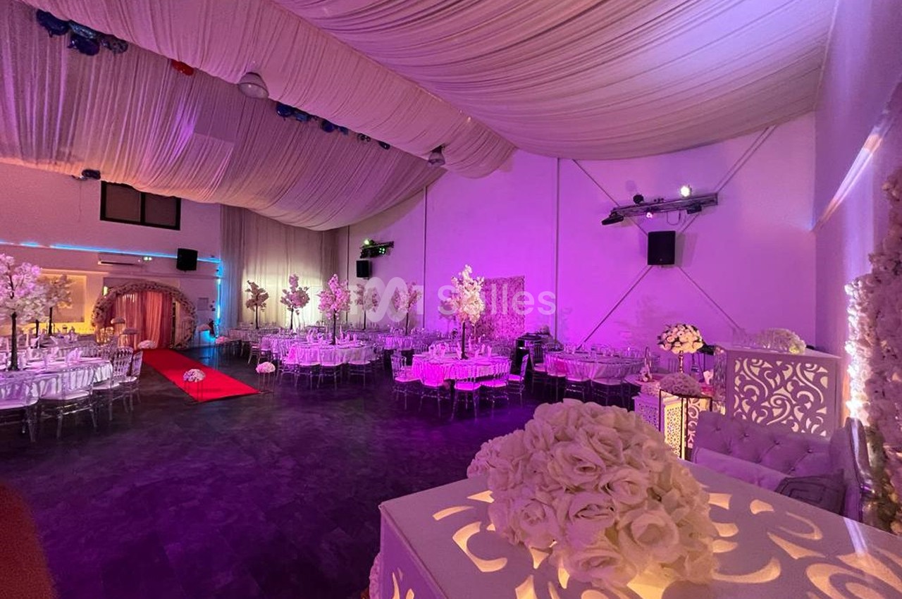 Salle de réception décorée avec des tables rondes, des fleurs et un éclairage rose sous un plafond drapé.