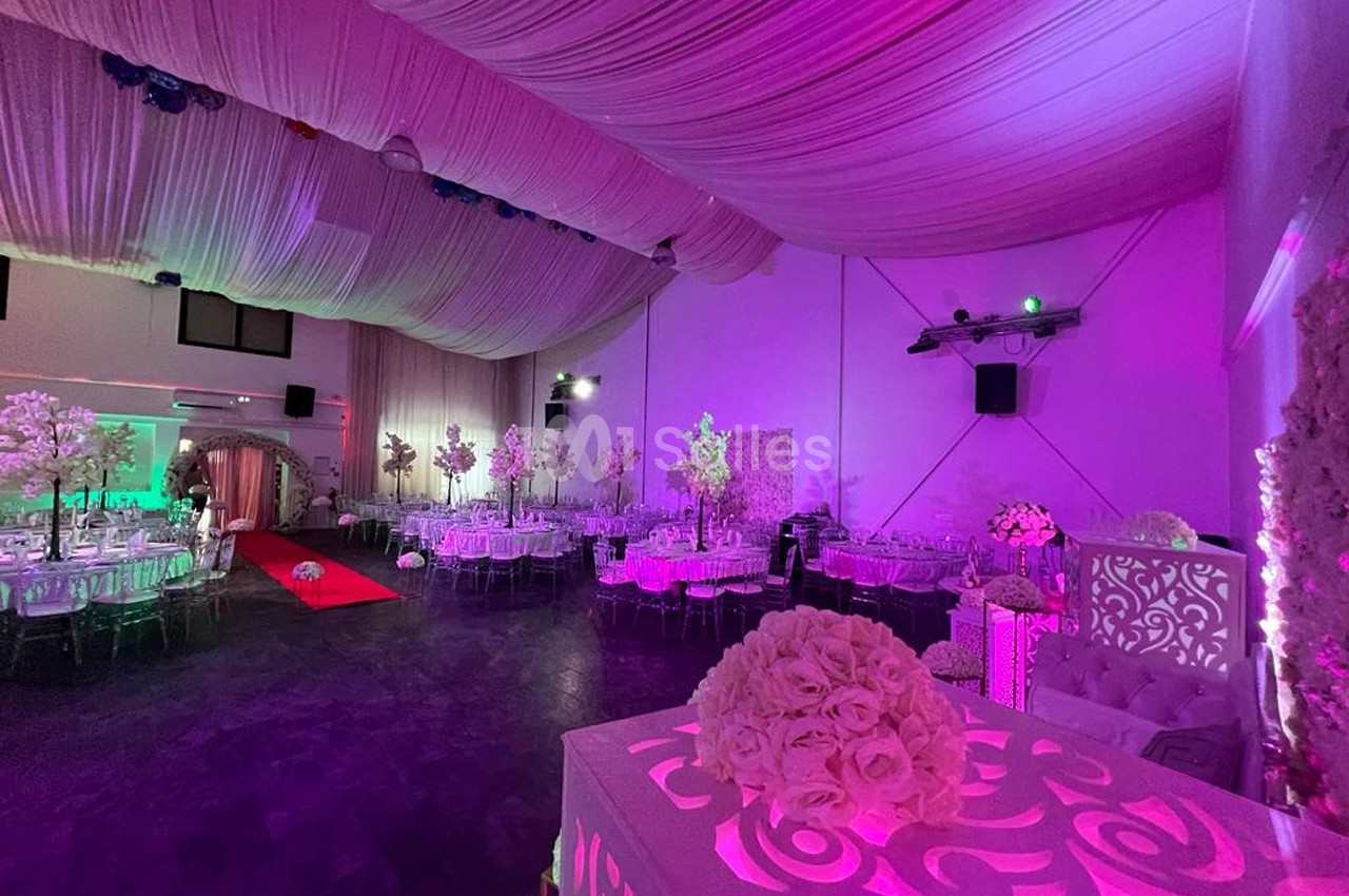 Salle de réception décorée avec des tables, des chaises transparentes, des fleurs blanches et un éclairage rose.