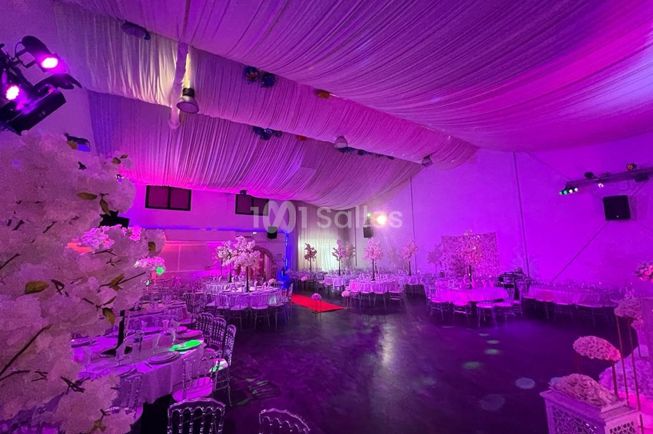 Salle de réception décorée avec des tables rondes, des chaises transparentes et un éclairage rose-violet.