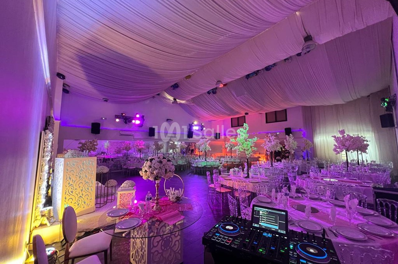 Salle de réception décorée avec des tables dressées, des fleurs blanches et un éclairage coloré, vue depuis une table de DJ.