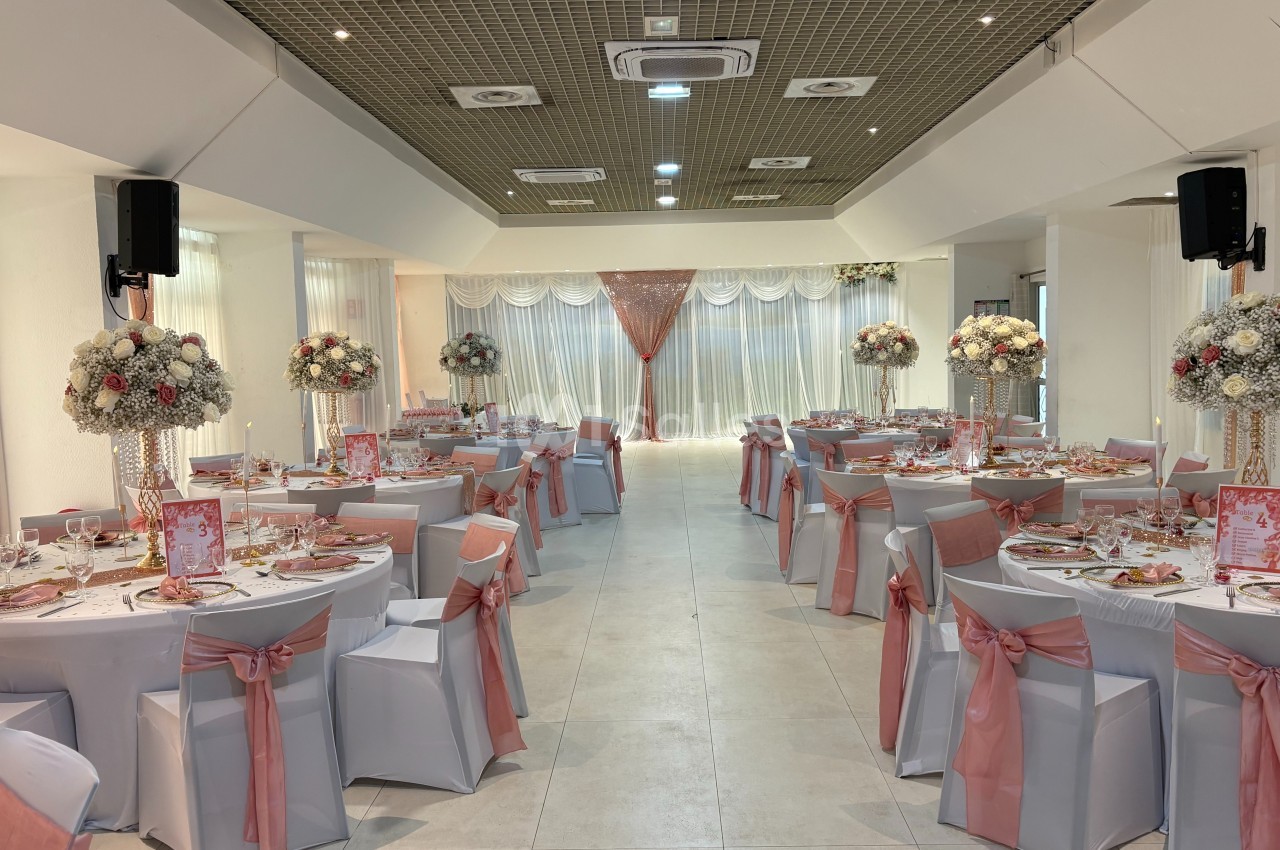 Salle de réception décorée pour un événement, avec tables rondes ornées de nappes blanches et rubans roses.