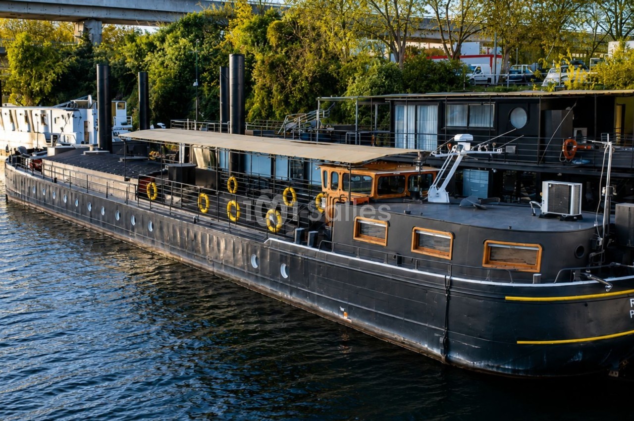 Bateau noir amarré sur une rivière, entouré de végétation et de structures urbaines en arrière-plan.