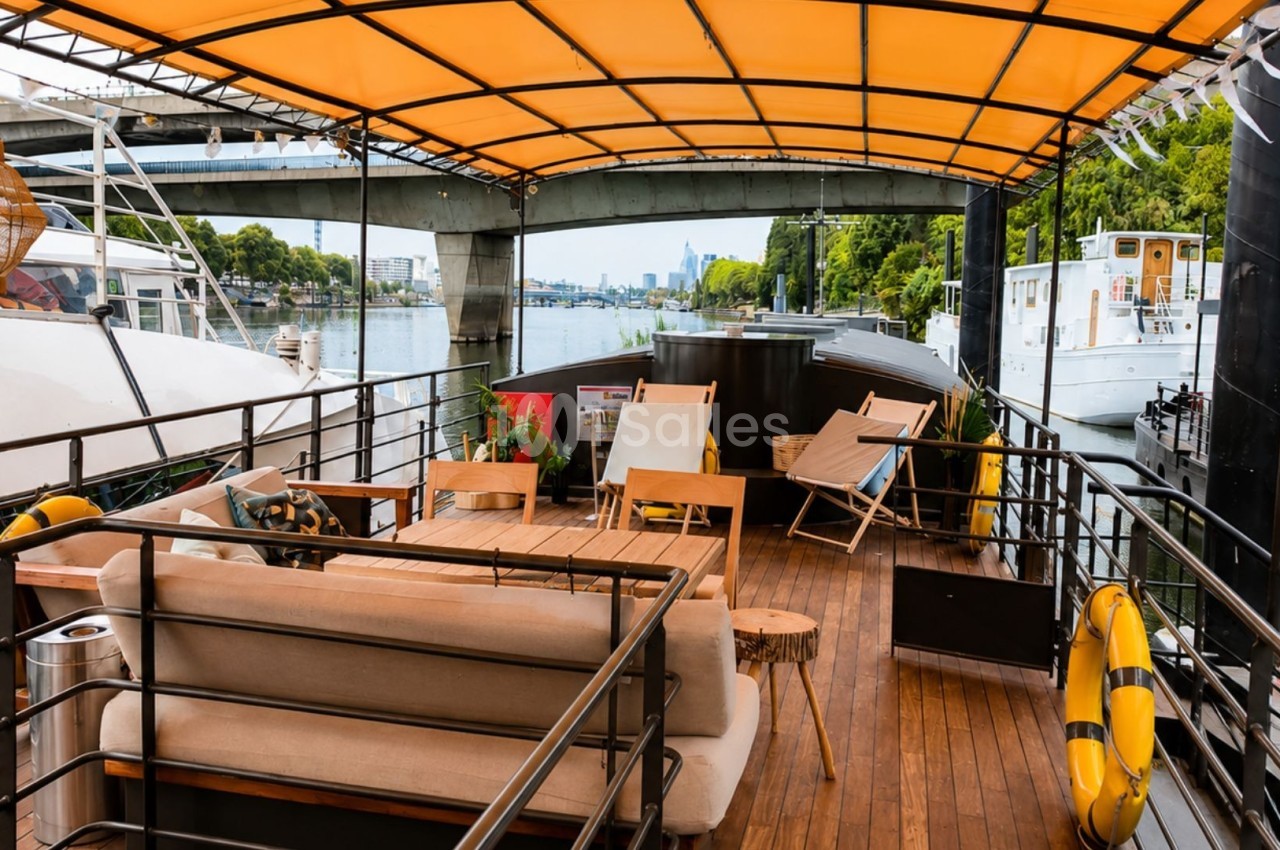 Terrasse en bois d'un bateau avec canapés, chaises longues et toit jaune, amarré près d'autres bateaux sur une rivière.