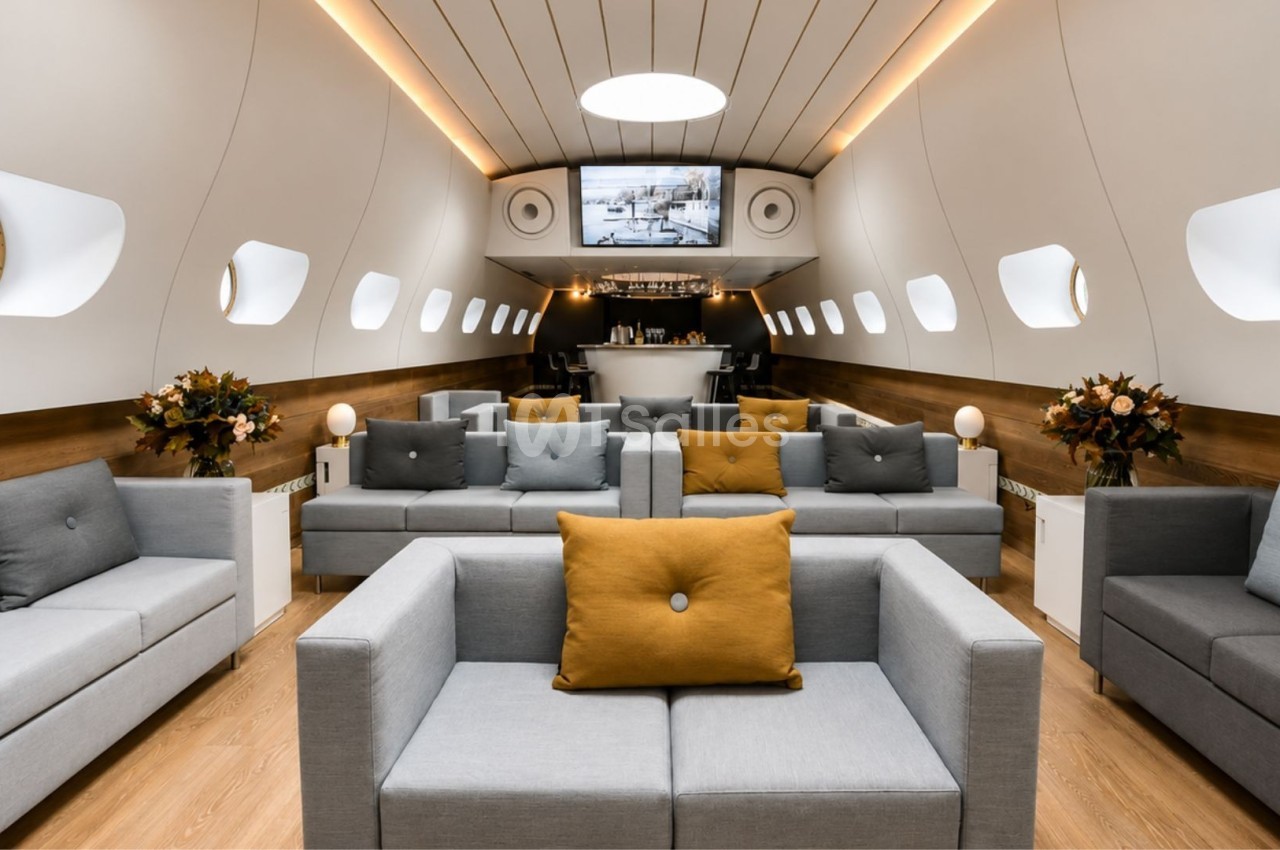 Intérieur d'un espace aménagé en style cabine d'avion avec canapés gris, coussins colorés et un bar au fond.