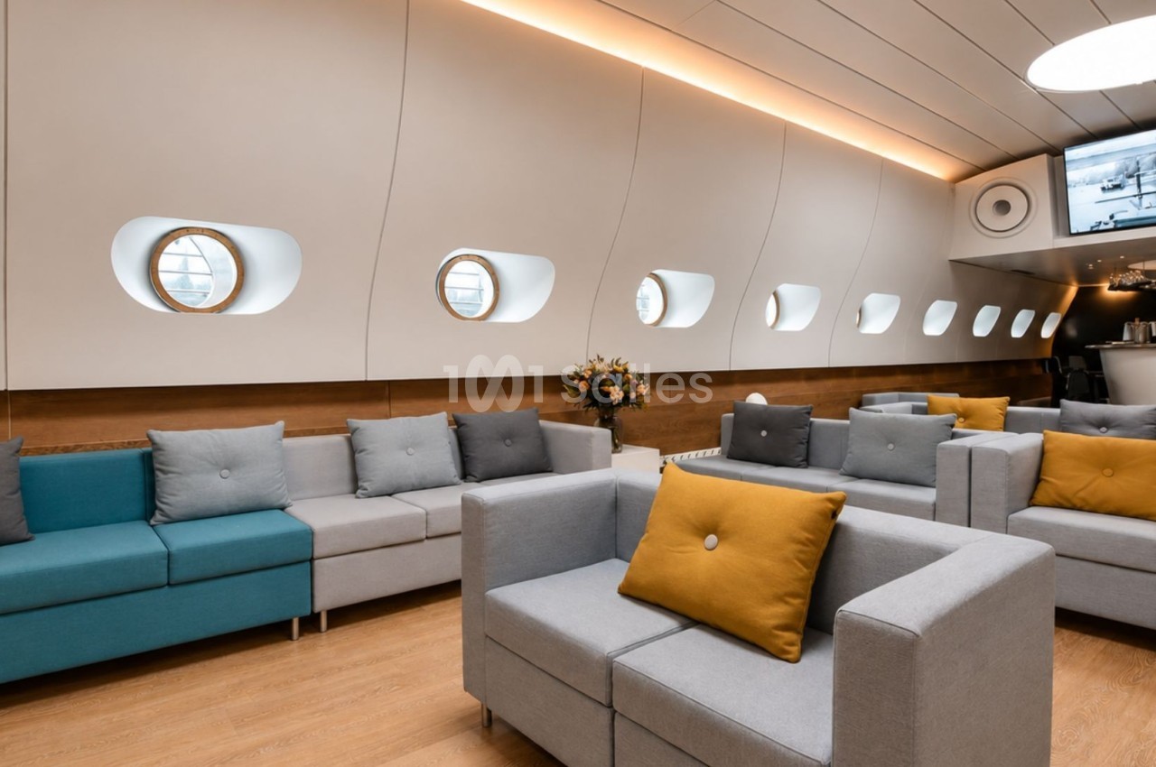 Salon moderne avec canapés colorés, éclairage chaleureux, hublots arrondis et écran mural au fond.