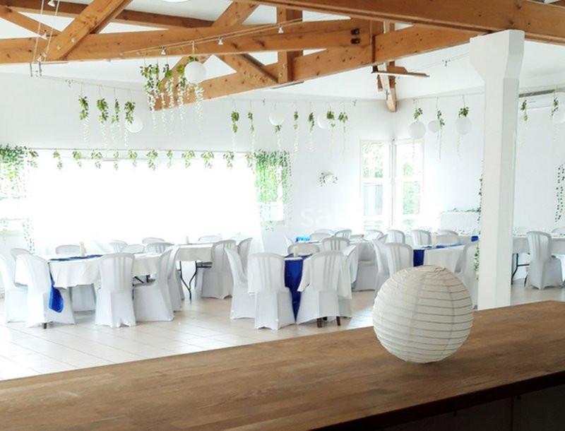 Salle lumineuse décorée avec des tables recouvertes de nappes blanches, ornées de touches bleues et de végétation suspendue.