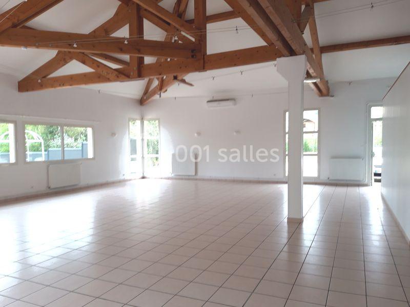 Salle lumineuse avec poutres apparentes, carrelage au sol, grandes fenêtres et murs blancs.