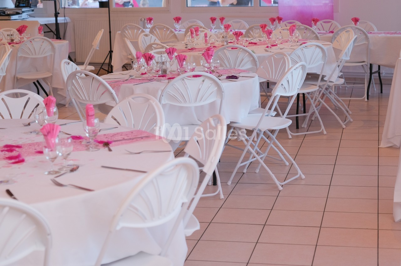 Salle décorée pour un événement, avec des tables rondes dressées, nappes blanches et accents roses.