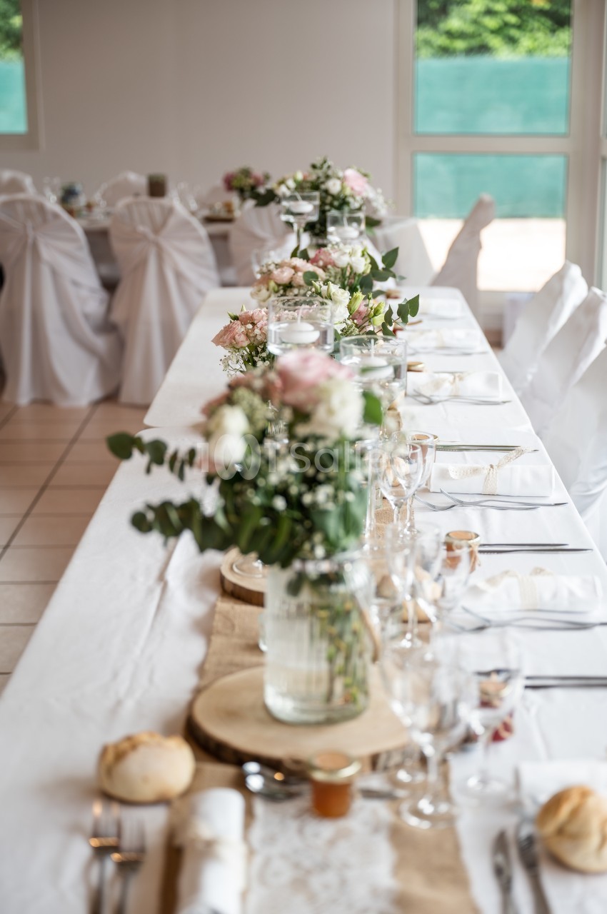 Table de réception décorée avec des fleurs, nappes blanches, vaisselle élégante et petits pains disposés.