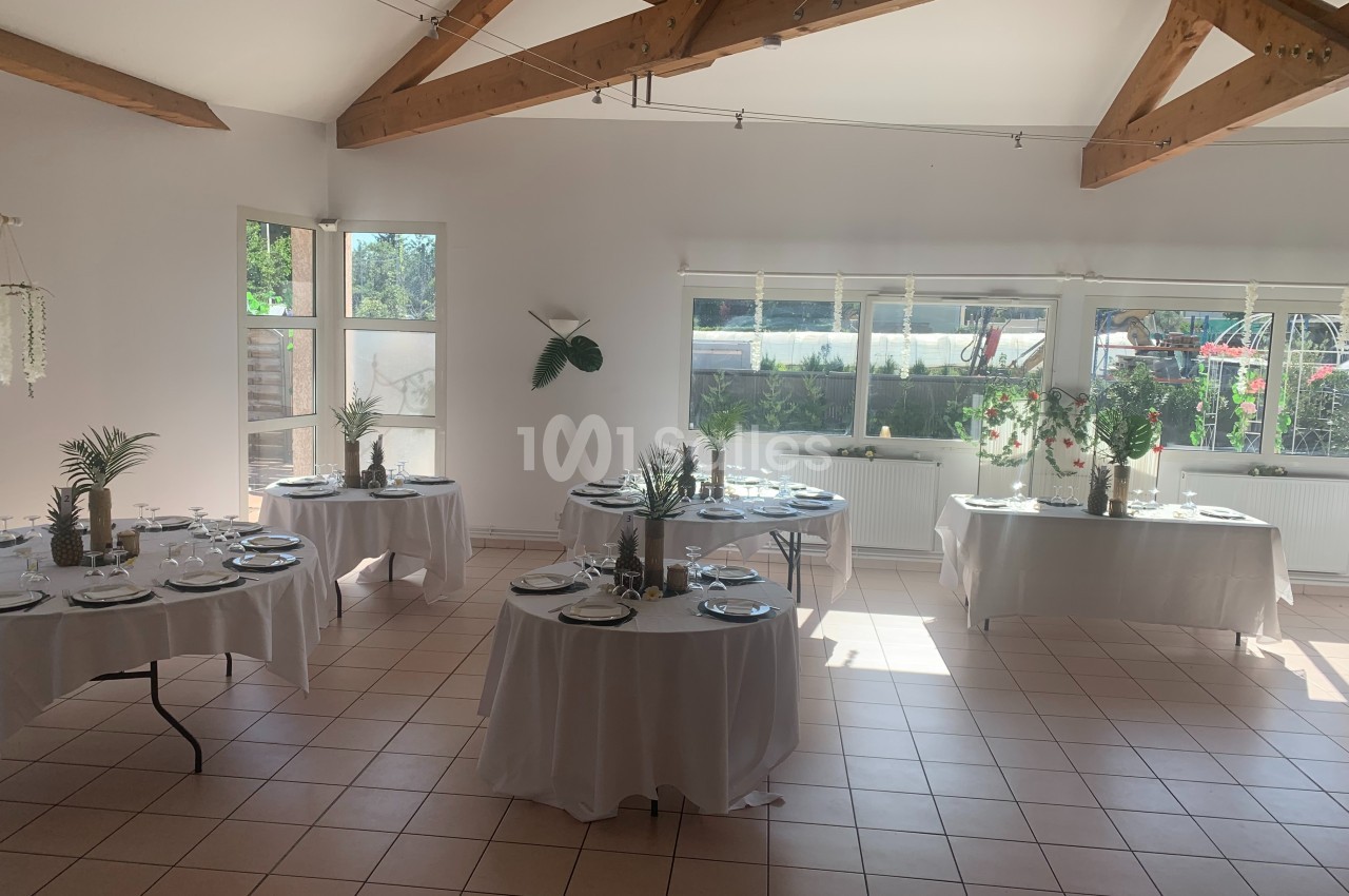 Salle lumineuse avec tables rondes dressées pour un repas, décorées de plantes et entourées de grandes fenêtres.
