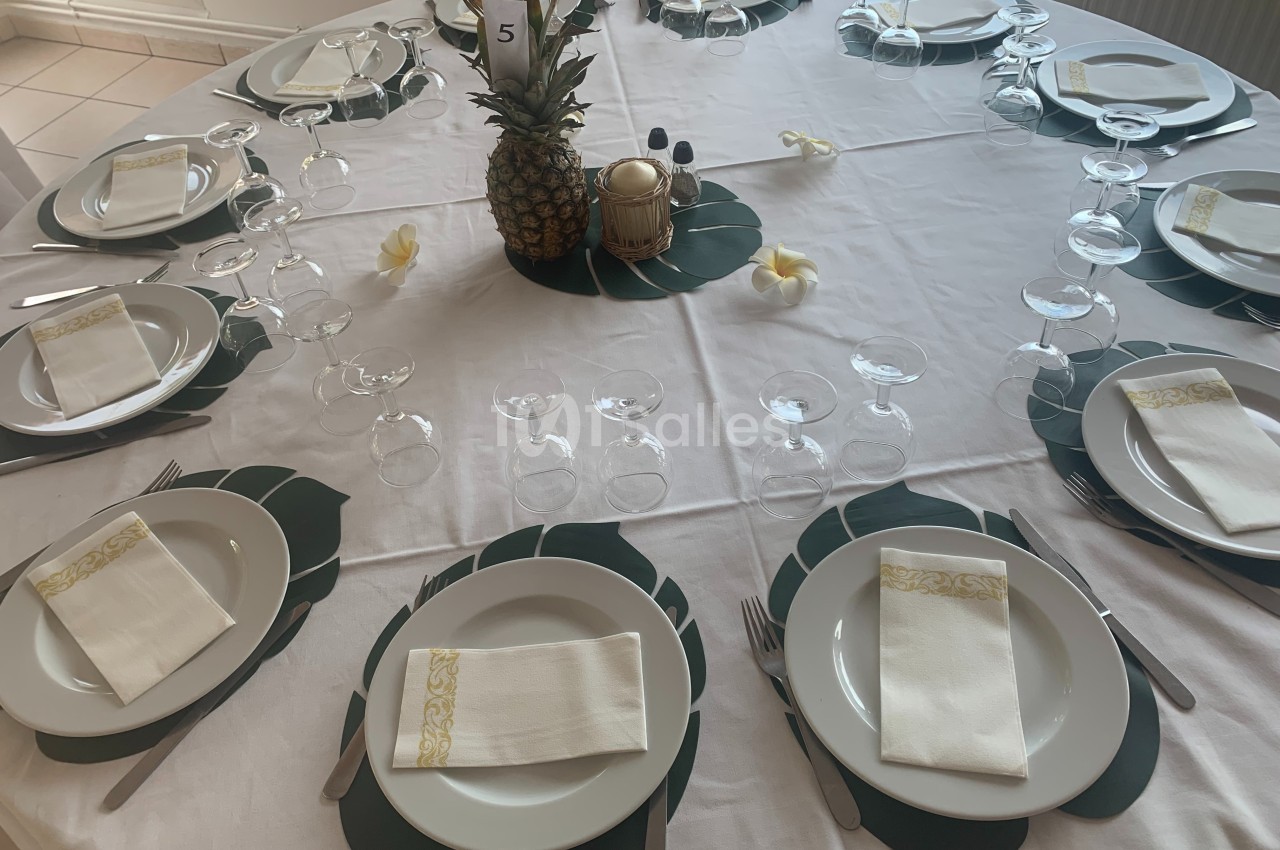 Table ronde dressée avec nappes blanches, assiettes, serviettes pliées, verres et décoration centrale avec un ananas.