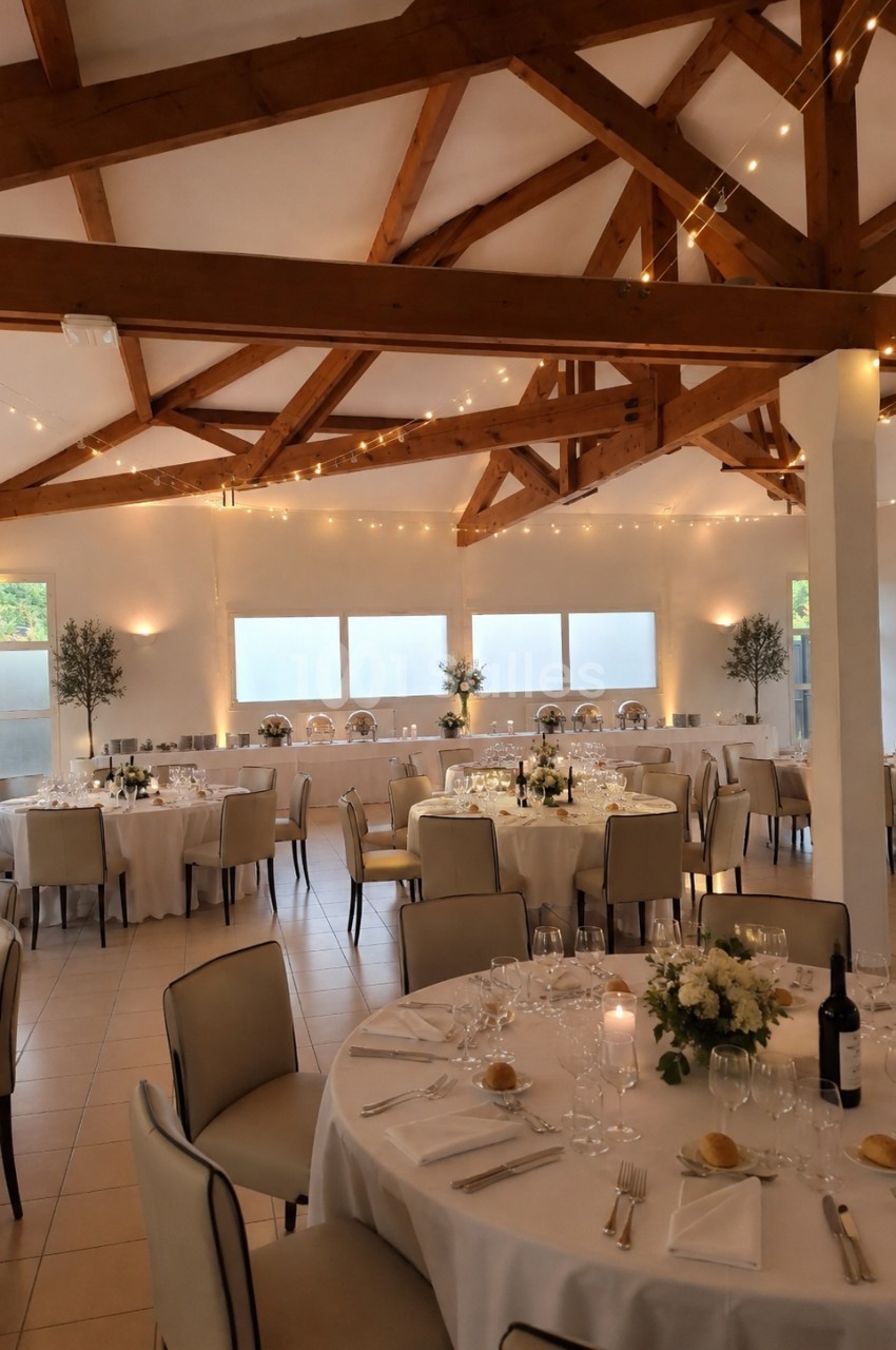 Salle de réception lumineuse avec tables rondes dressées, décorée de guirlandes lumineuses et fleurs blanches.