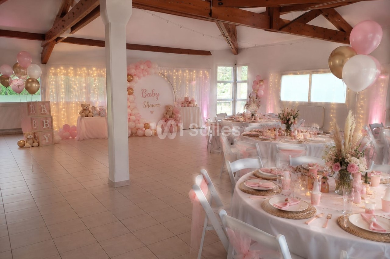 Salle décorée pour une baby shower avec tables dressées, ballons roses et dorés, et guirlandes lumineuses.
