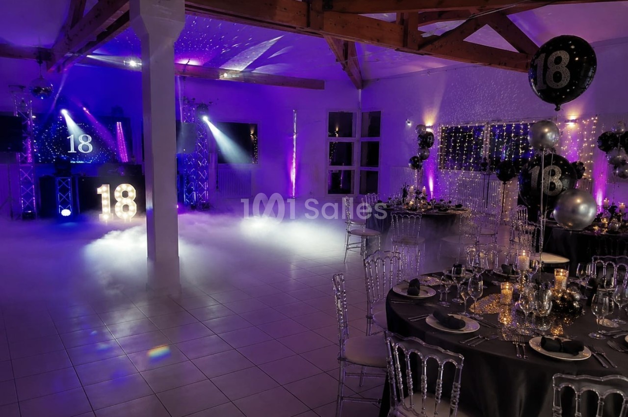 Salle décorée pour une fête d'anniversaire avec tables dressées, ballons ’18’ et éclairages violets.