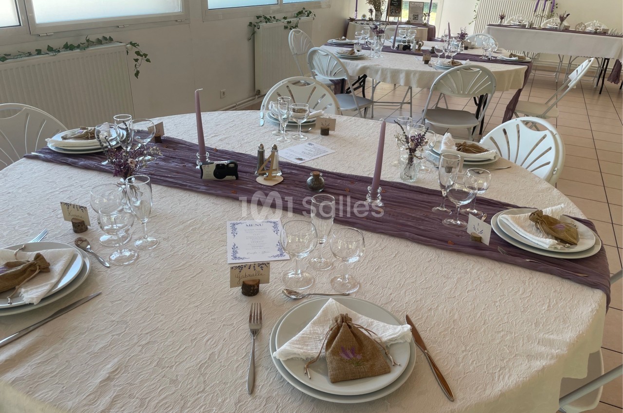 Table décorée pour un repas, avec nappes blanches, chemins de table violets, vaisselle et petits bouquets.