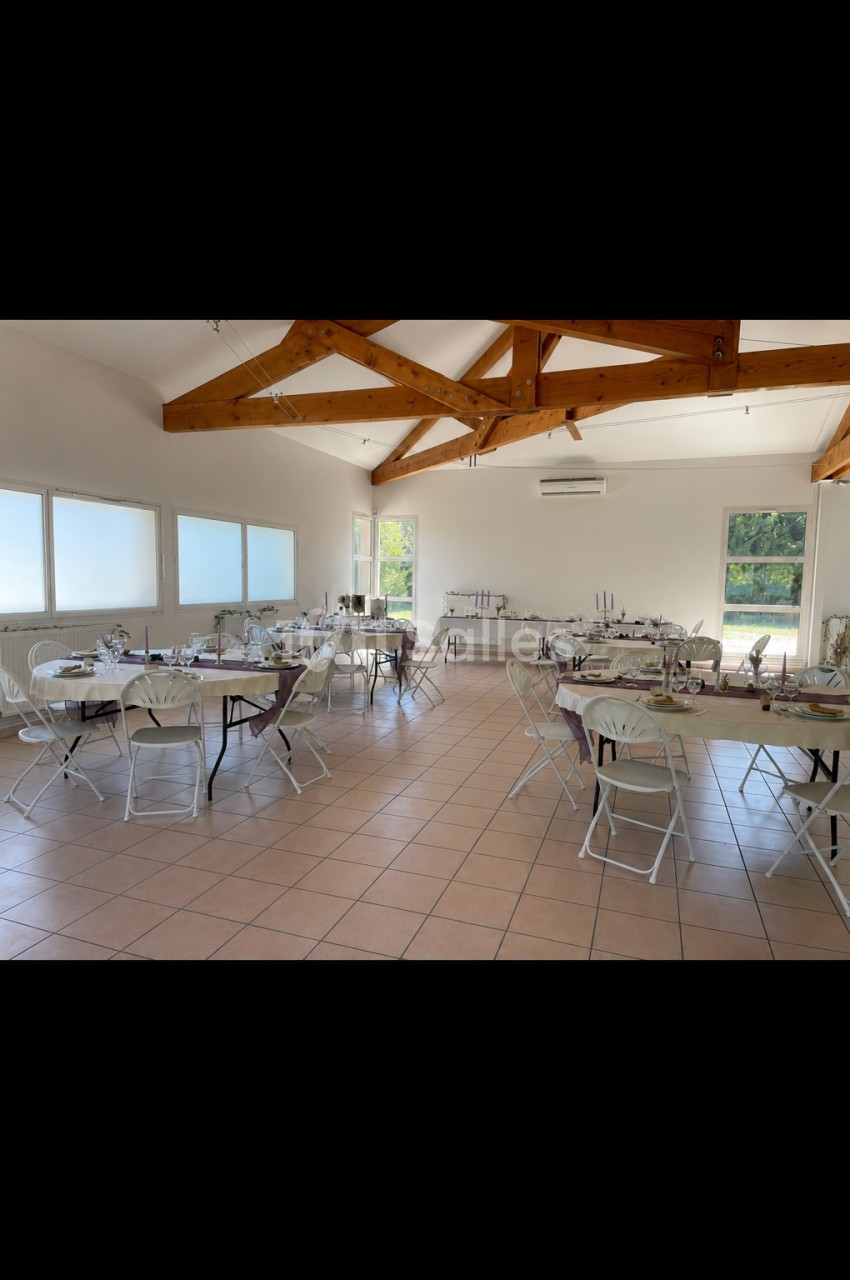 Salle lumineuse avec tables rondes dressées, chaises pliantes blanches et poutres apparentes au plafond.