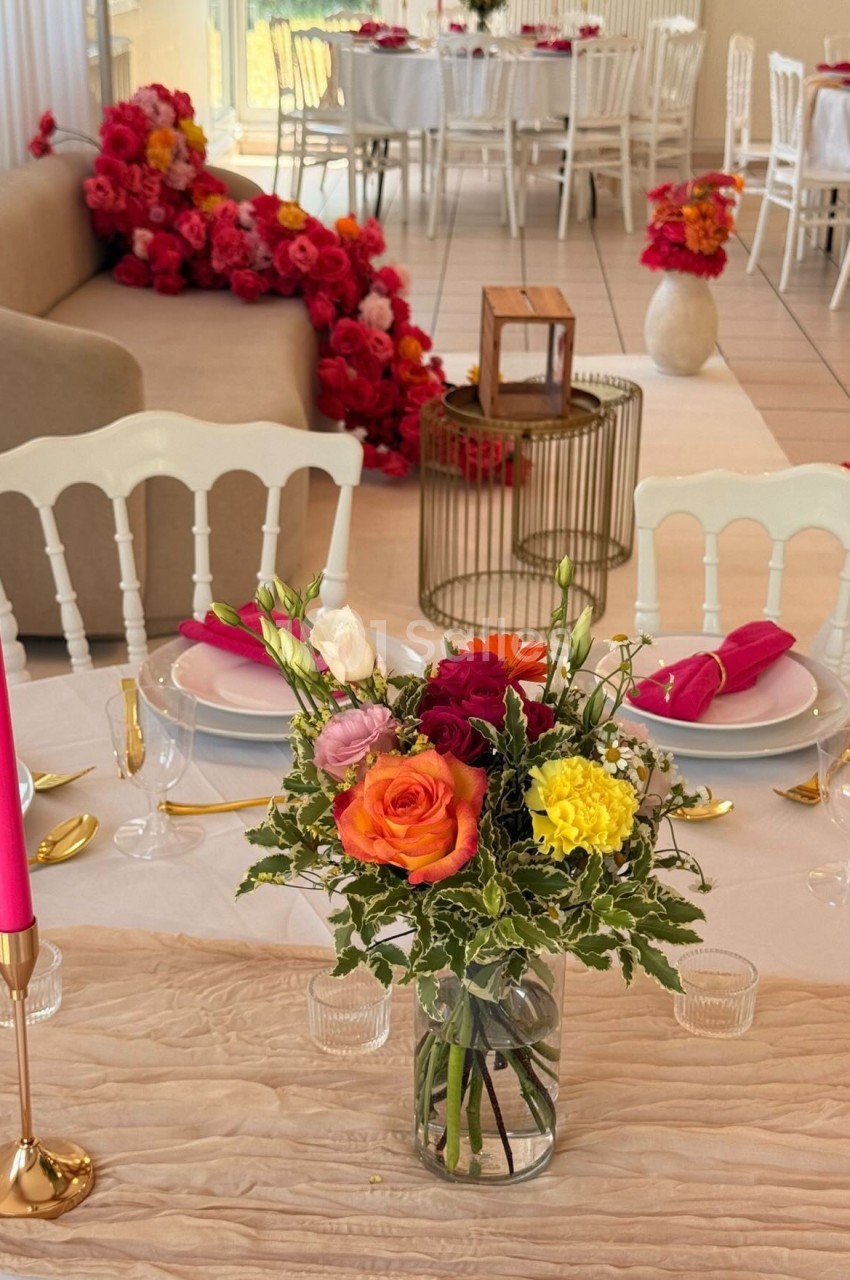 Table décorée avec un bouquet de fleurs colorées, bougie rose et vaisselle élégante, dans une salle ornée de roses rouges.