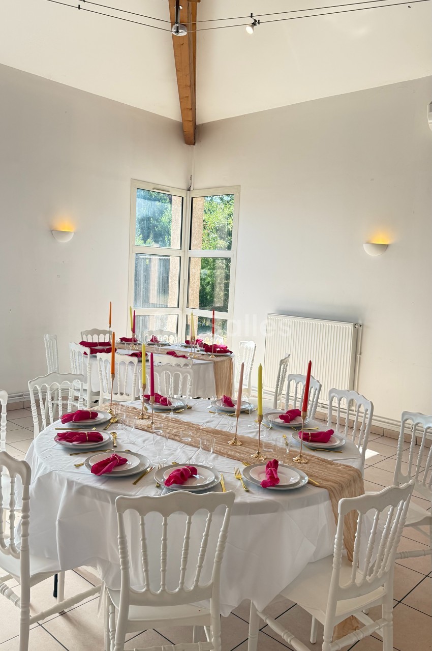 Salle lumineuse avec tables décorées de nappes blanches, bougies colorées et serviettes rouges, prête pour un repas.