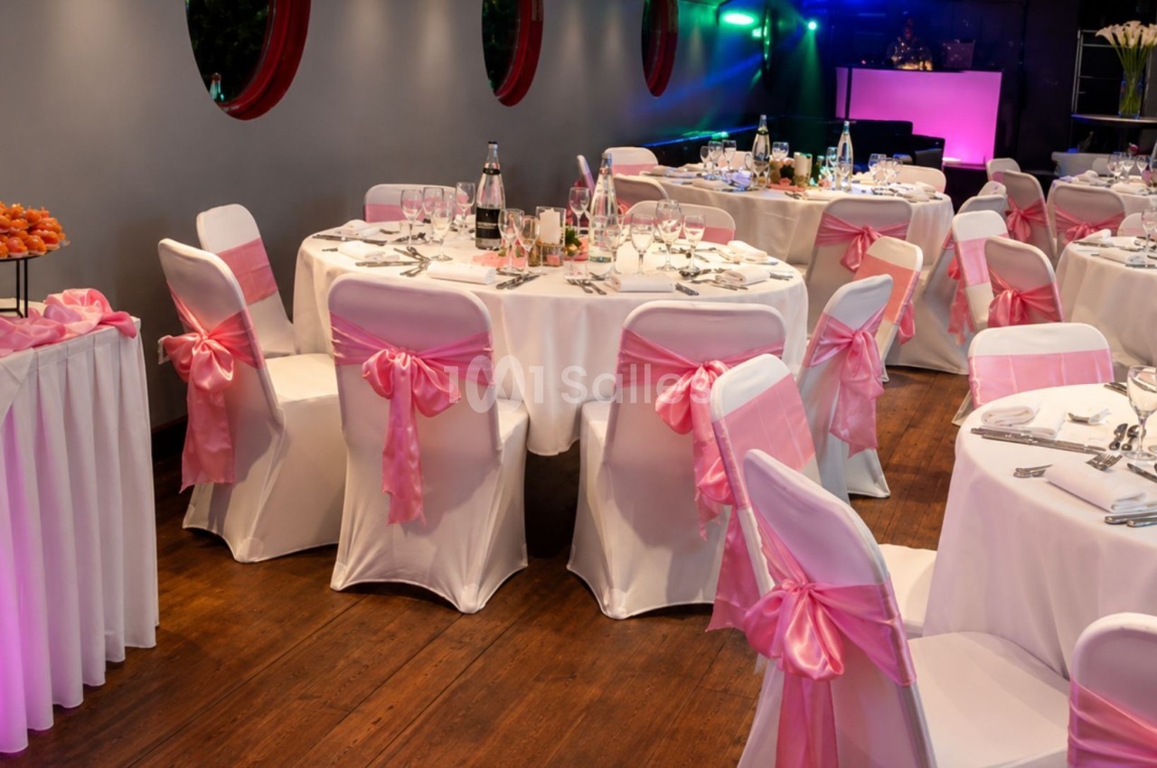 Salle de réception décorée avec des tables rondes, nappes blanches et chaises ornées de nœuds roses.