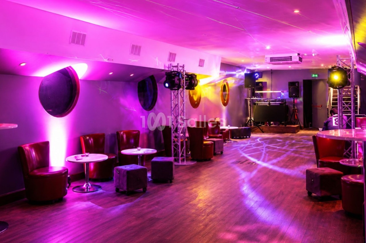Salle de réception avec éclairage violet, tables rondes, fauteuils rouges et piste de danse équipée de matériel audio.