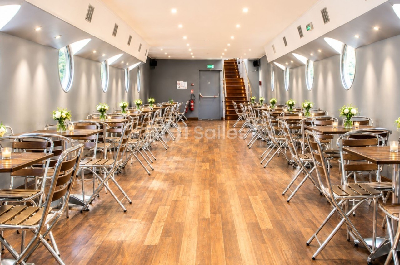 Salle lumineuse avec parquet, tables et chaises en métal, décorée de bouquets, et fenêtres rondes sur les murs.