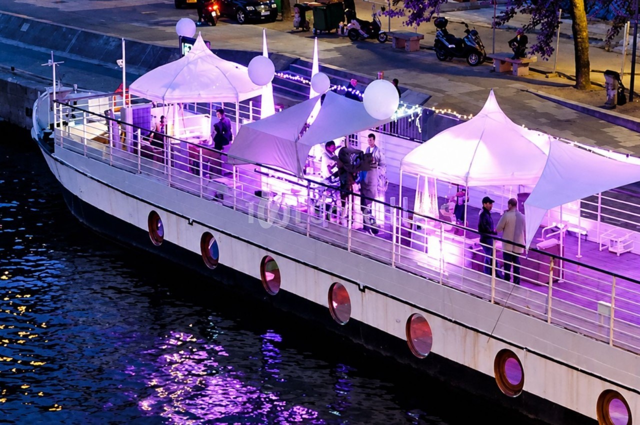 Bateau amarré avec des tentes blanches éclairées en violet, accueillant des personnes sur le pont en soirée.