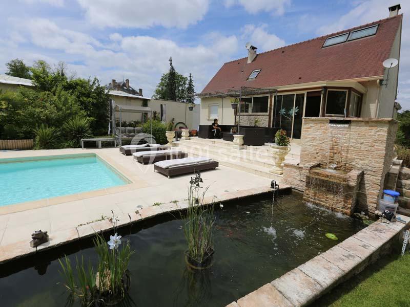 Maison avec terrasse, piscine, bassin ornemental et jardin arboré sous un ciel partiellement nuageux.