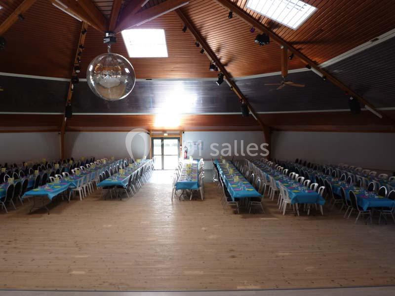 Salle spacieuse avec plafond en bois, tables alignées et dressées avec nappes bleues, éclairée par des fenêtres et un lustre…