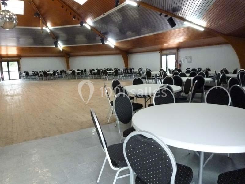 Salle spacieuse avec parquet, tables rondes blanches et chaises noires, éclairée par des lumières au plafond en bois.
